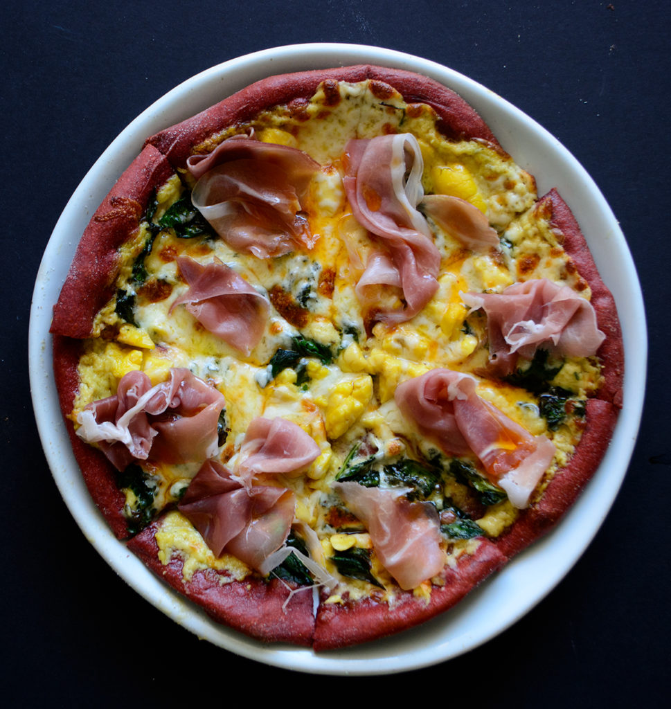 Beetroot-pizza-dough-topped-with-rich-ackee-cream,-ackees,-mozza-and-marinated-greens,-finished-with-proscuitto-raw-honey-and-chilli-oil